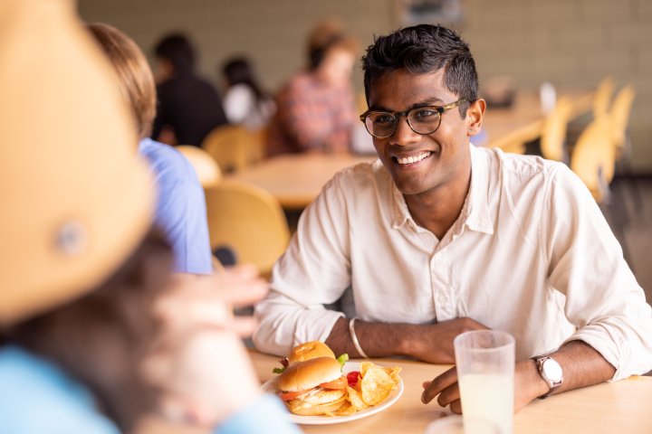 A student wearing glasses smiles while sitting at a dining hall table. In front of them is a plate with a sandwich, chips, and a glass of lemonade. The background shows other students seated at tables, blurred, creating a casual and friendly atmosphere.